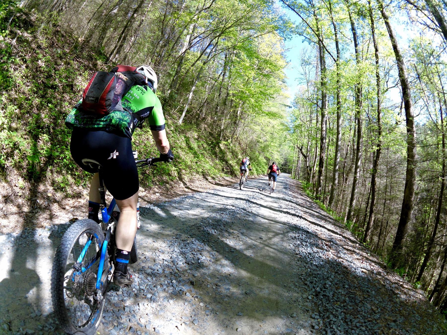 A group of three mountain bikers riding on a gravel path surrounded by lush green trees. The first cyclist is wearing a green jersey with black shorts and a red backpack, while the other two cyclists are visible in the background on the same trail. The scene captures a sunny day with blue skies and fresh foliage. Bear Creek mountain bike trail.