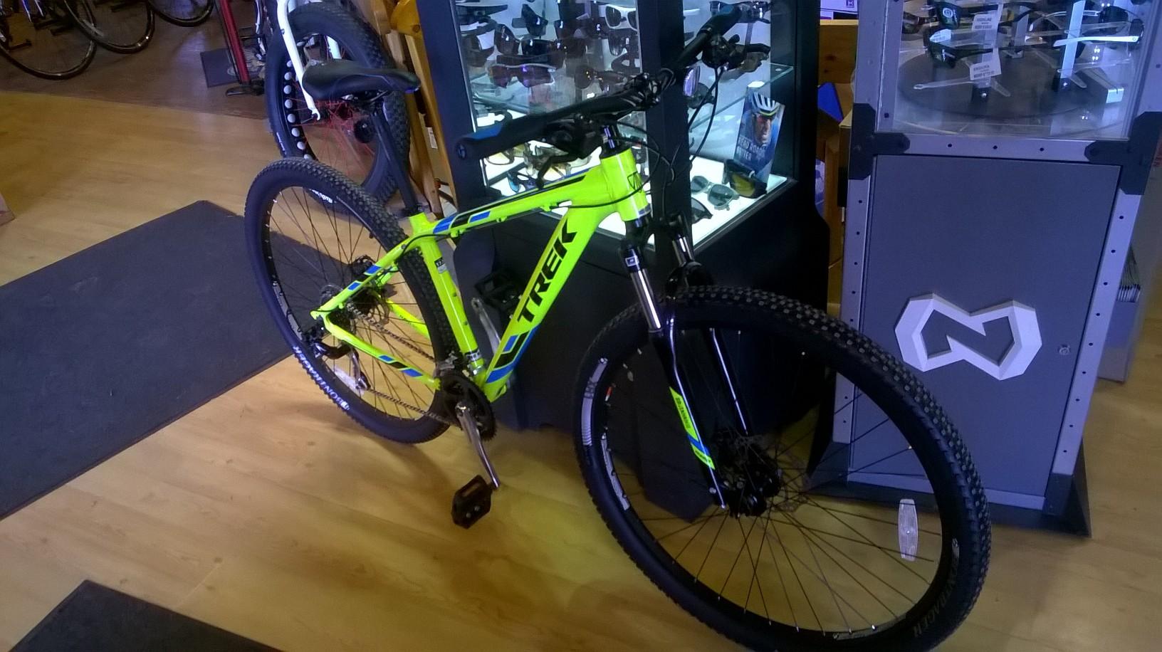 Trek Marlin 5: A bright yellow and blue mountain bike displayed inside a bike shop, with a glass cabinet in the background showcasing cycling accessories and sunglasses. The bike features thick tires and a sturdy frame, positioned on a wooden floor with various biking equipment visible in the store.