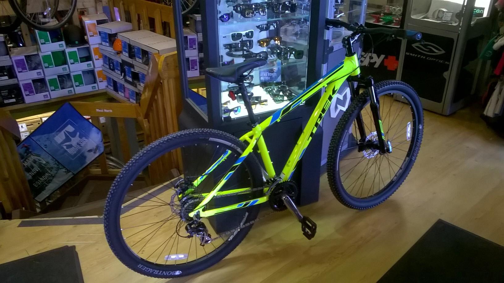 Trek Marlin 5: Alt tag: A bright green and blue mountain bike displayed inside a retail store, with sunglasses and other merchandise visible in the background.