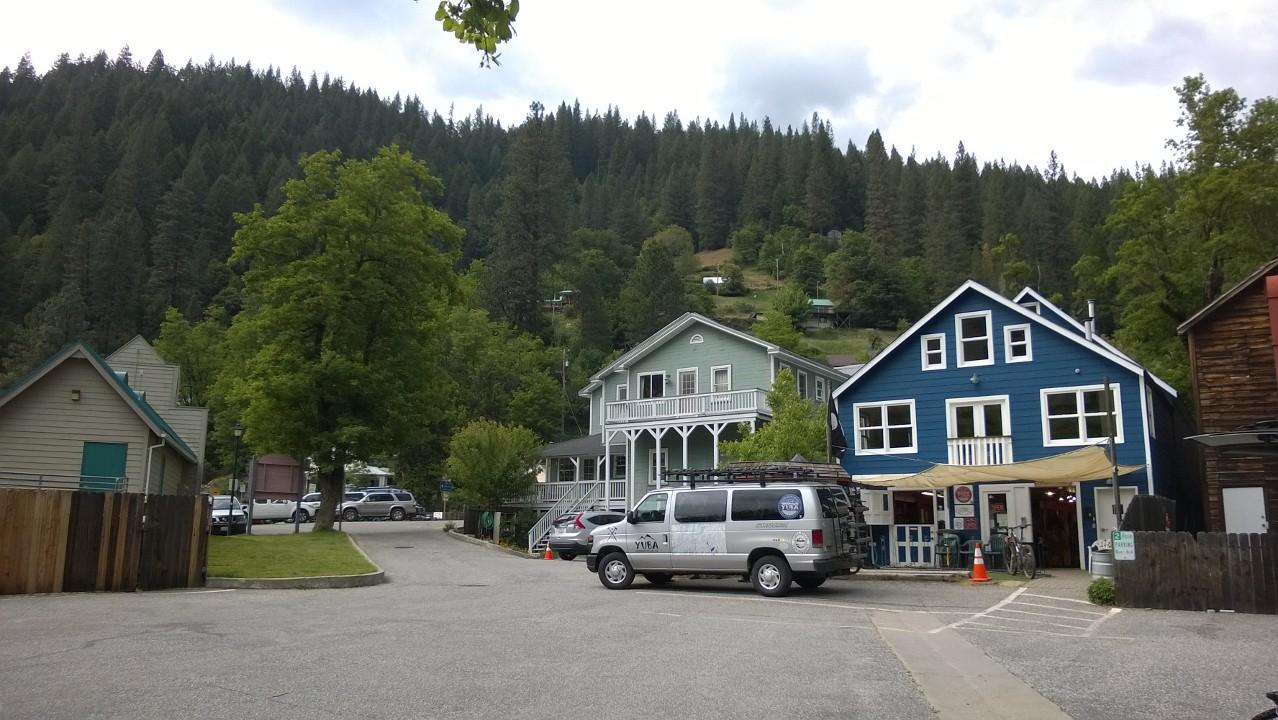 A scenic view of a quaint small town featuring a mix of colorful buildings, including a blue house and a light green structure, set against a backdrop of lush forested hills. Parked vehicles line a small road, with trees and greenery enhancing the charming atmosphere. Cloudy skies add a soft light to the scene.