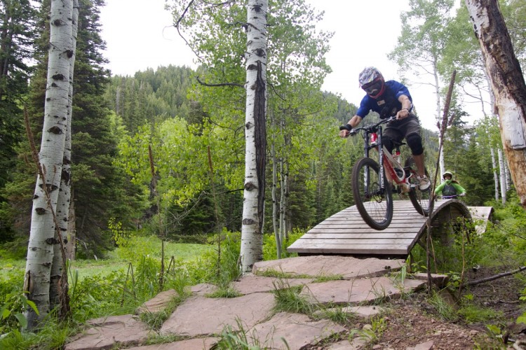 A mountain biker in protective gear catching air over a wooden ramp on a trail surrounded by trees and greenery, with another cyclist visible in the background.