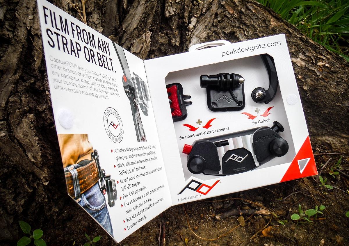 Peak Design Capture P.O.V. Action Mount: A product packaging for the CapturePOV by Peak Design, designed for mounting action cameras and point-and-shoot cameras. The image shows the inside of the box with various mounting accessories displayed, including clips and adapters. The background features a tree trunk and grass, emphasizing an outdoor setting. Text on the packaging highlights the product's versatility, usability with straps or belts, and compatibility with various camera types.