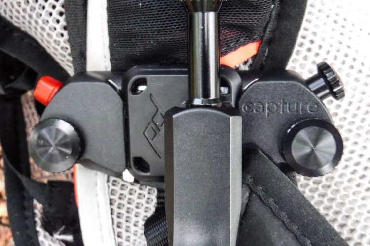 Close-up of a camera clip attachment mounted on a backpack, featuring adjustable knobs and a sleek black design, showcasing a practical tool for outdoor photography. The background includes textured mesh fabric of the backpack.