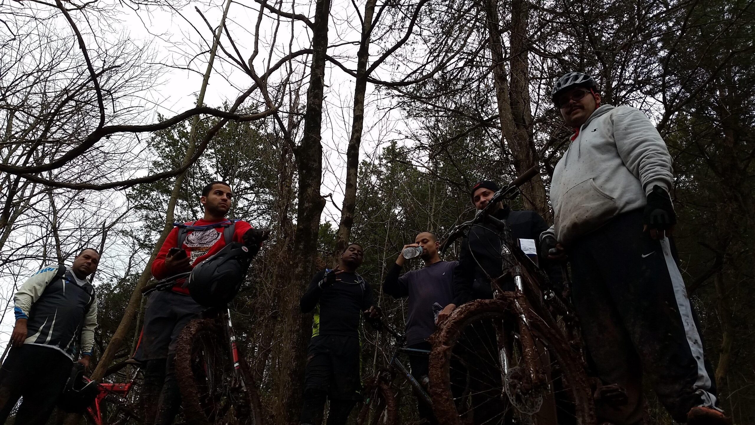 Group of six mountain bikers standing near their bicycles in a wooded area. They are dressed in casual sportswear, with some showing mud on their clothes, indicating a recent ride. The trees are bare, and the sky appears overcast, suggesting a cloudy day. One rider is drinking water, while others are posing or looking at the camera. Six Mile Run mountain bike trail.