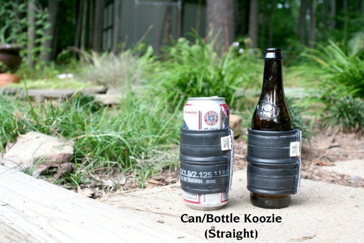 Image of a can and a bottle, both insulated with black koozies, placed on a wooden surface in a garden setting. The can is positioned on the left and the bottle on the right, with grass and trees in the background. The koozies are designed to keep drinks cool.