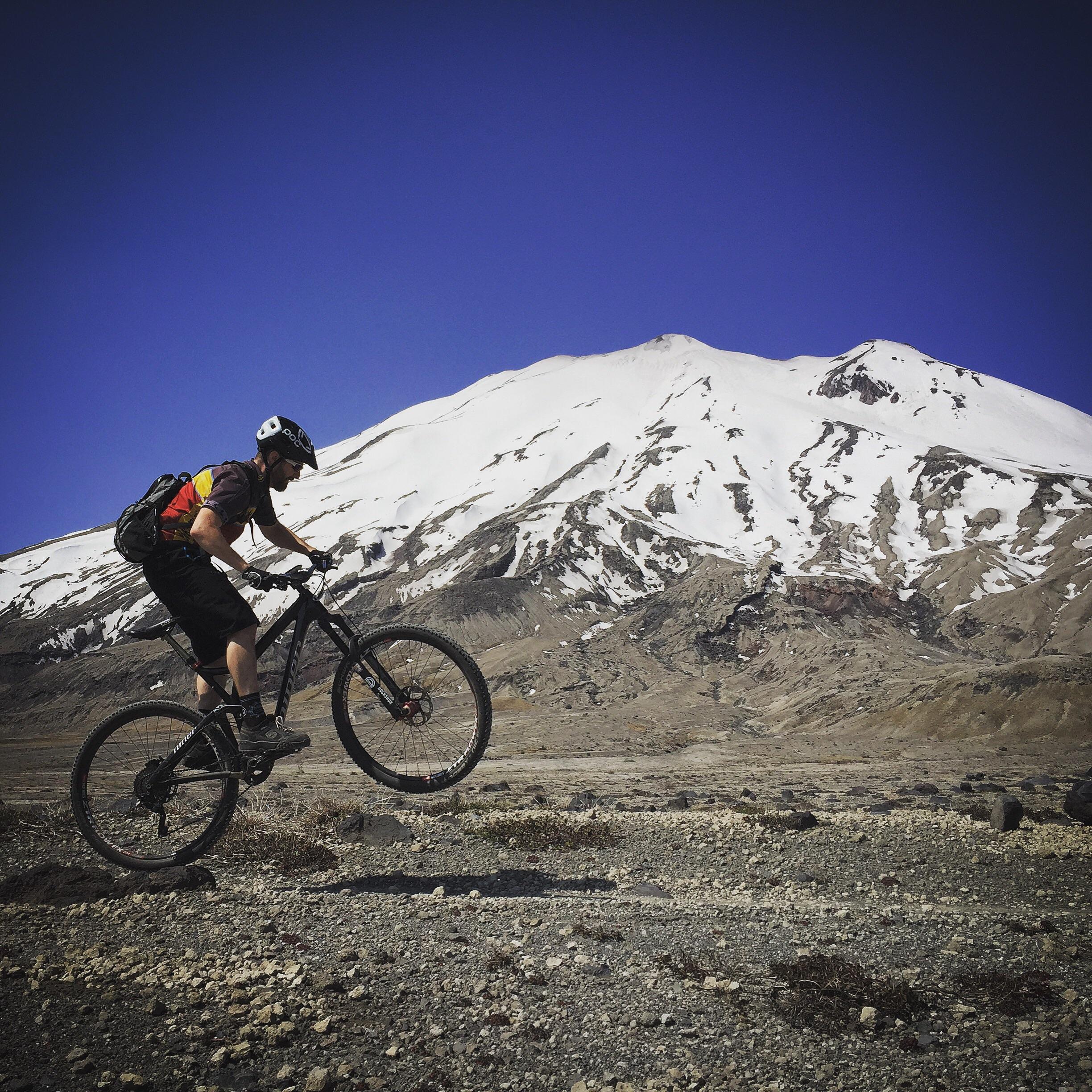 A mountain biker performing a wheelie on rocky terrain with a snow-capped mountain and clear blue sky in the background. The rider is wearing a helmet and a backpack, showcasing an adventurous outdoor scene. Ape Canyon#234, Abraham#216d, Smith Creek#225 Trails mountain bike trail.
