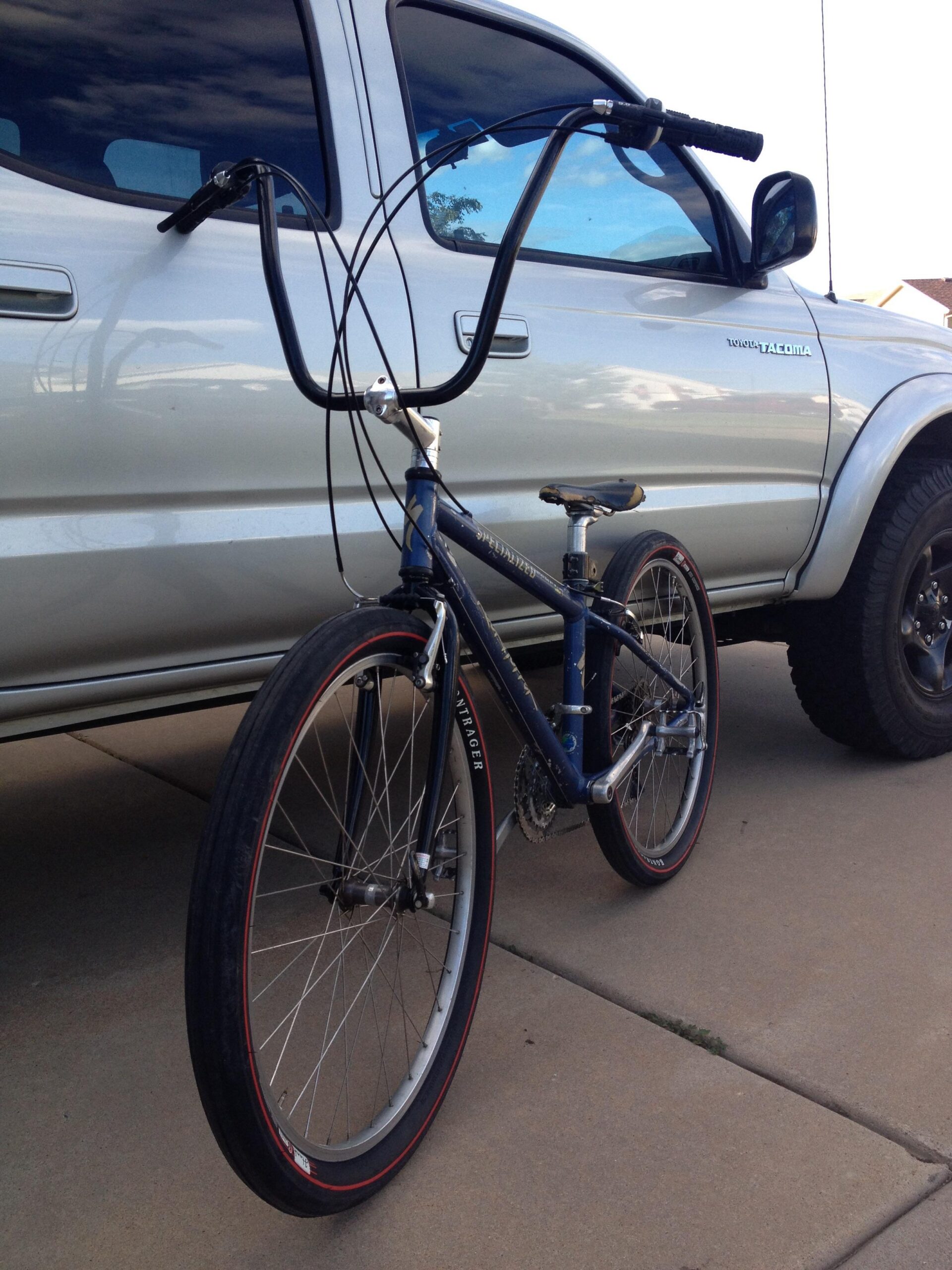 Specialized Rockhopper: A blue bicycle with high handlebars is parked on a concrete driveway next to a silver pickup truck. The bike features large wheels with red accents and a simple frame design, showcasing a unique vintage style. The truck's window reflects the sky and surroundings, adding depth to the image.