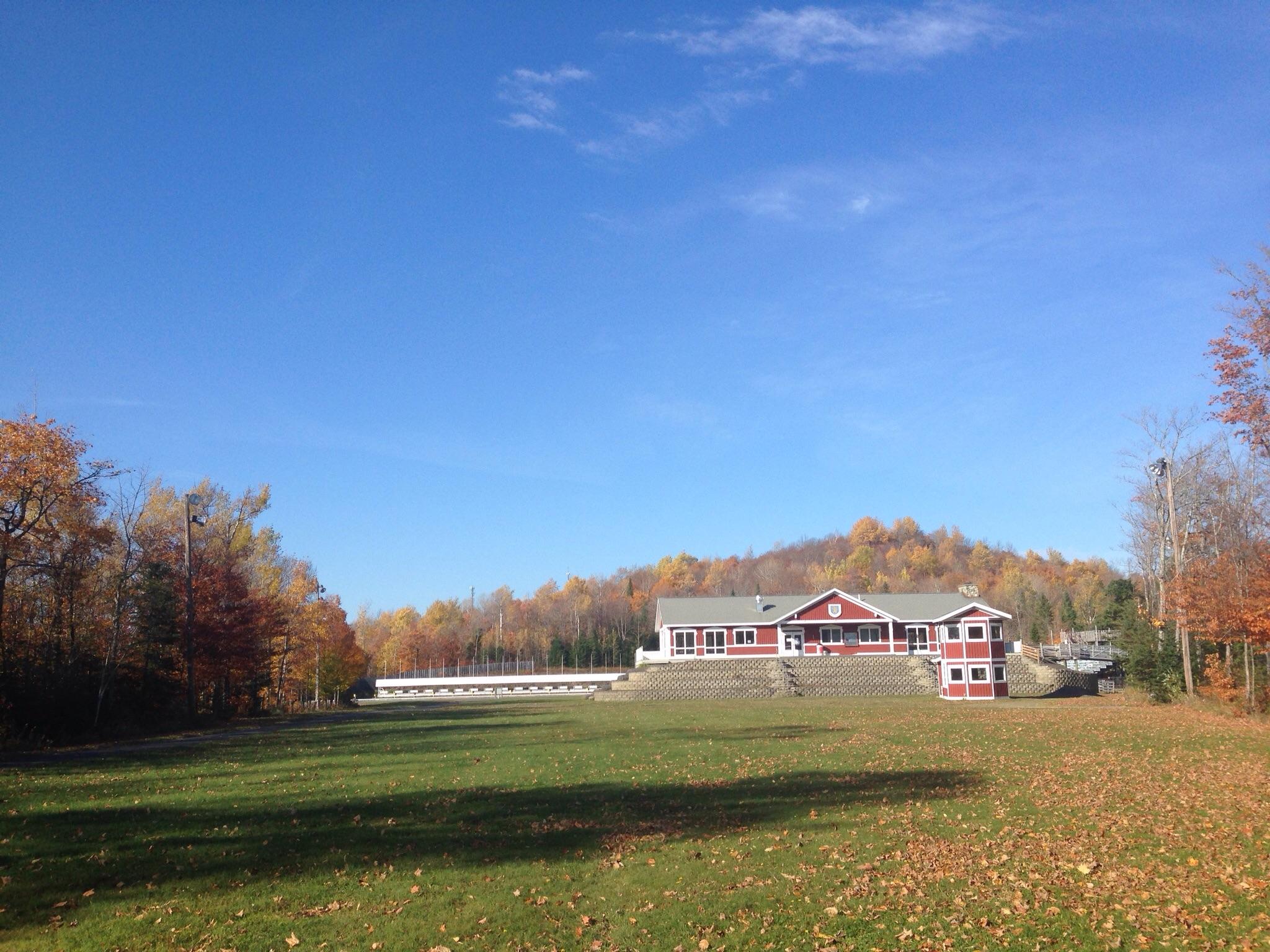 A scenic view of a spacious green field in autumn, featuring a red building on the right side with a gray roof. In the background, colorful autumn trees line the hillside, and a clear blue sky enhances the tranquil atmosphere. Nordic Heritage Center mountain bike trail.