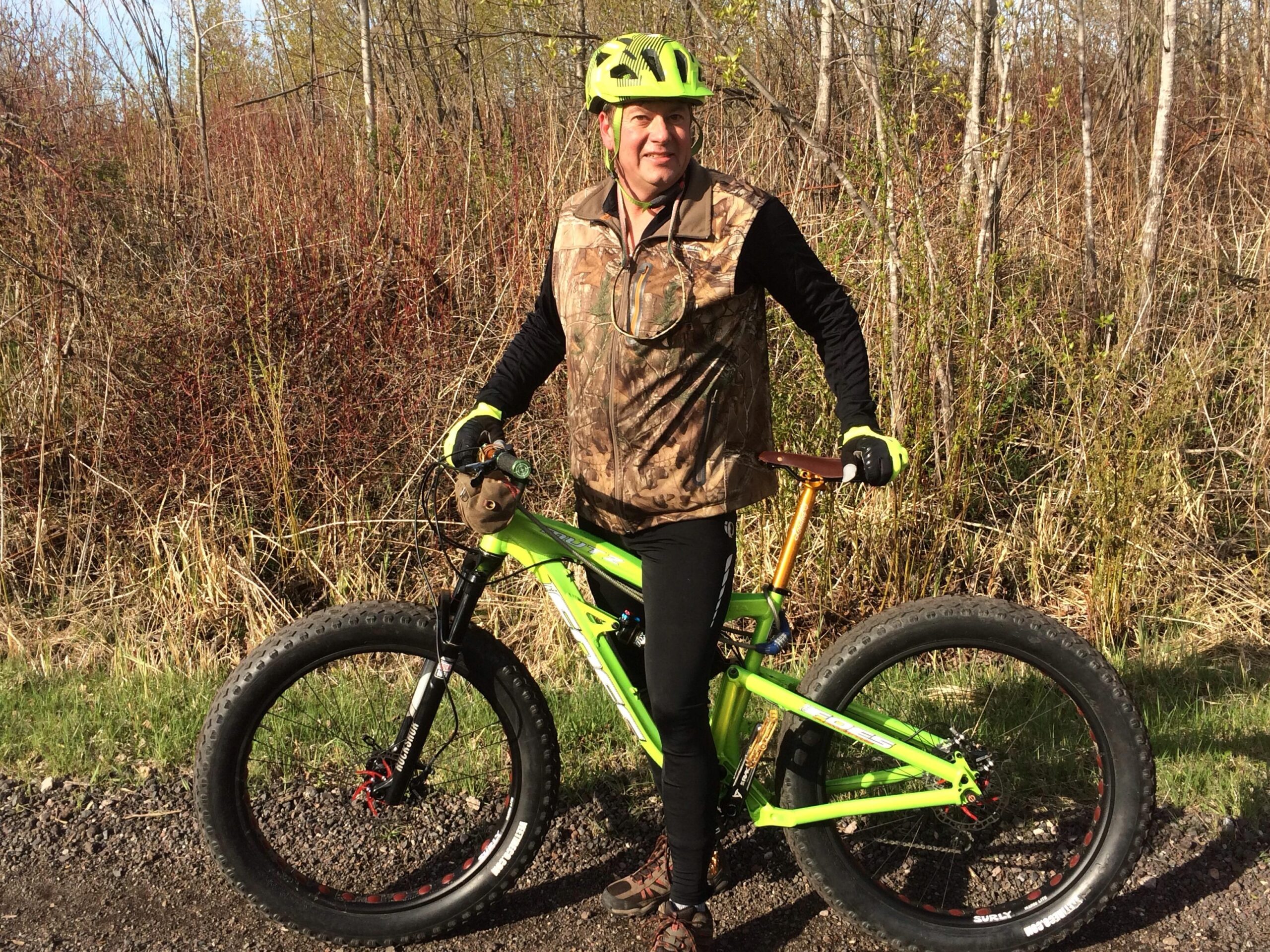 Foes Mutz: A person standing beside a bright green fat bike on a gravel path, wearing a camouflage vest, black leggings, and a bright yellow helmet and gloves. The background features greenery and bushes, indicating a natural outdoor setting.