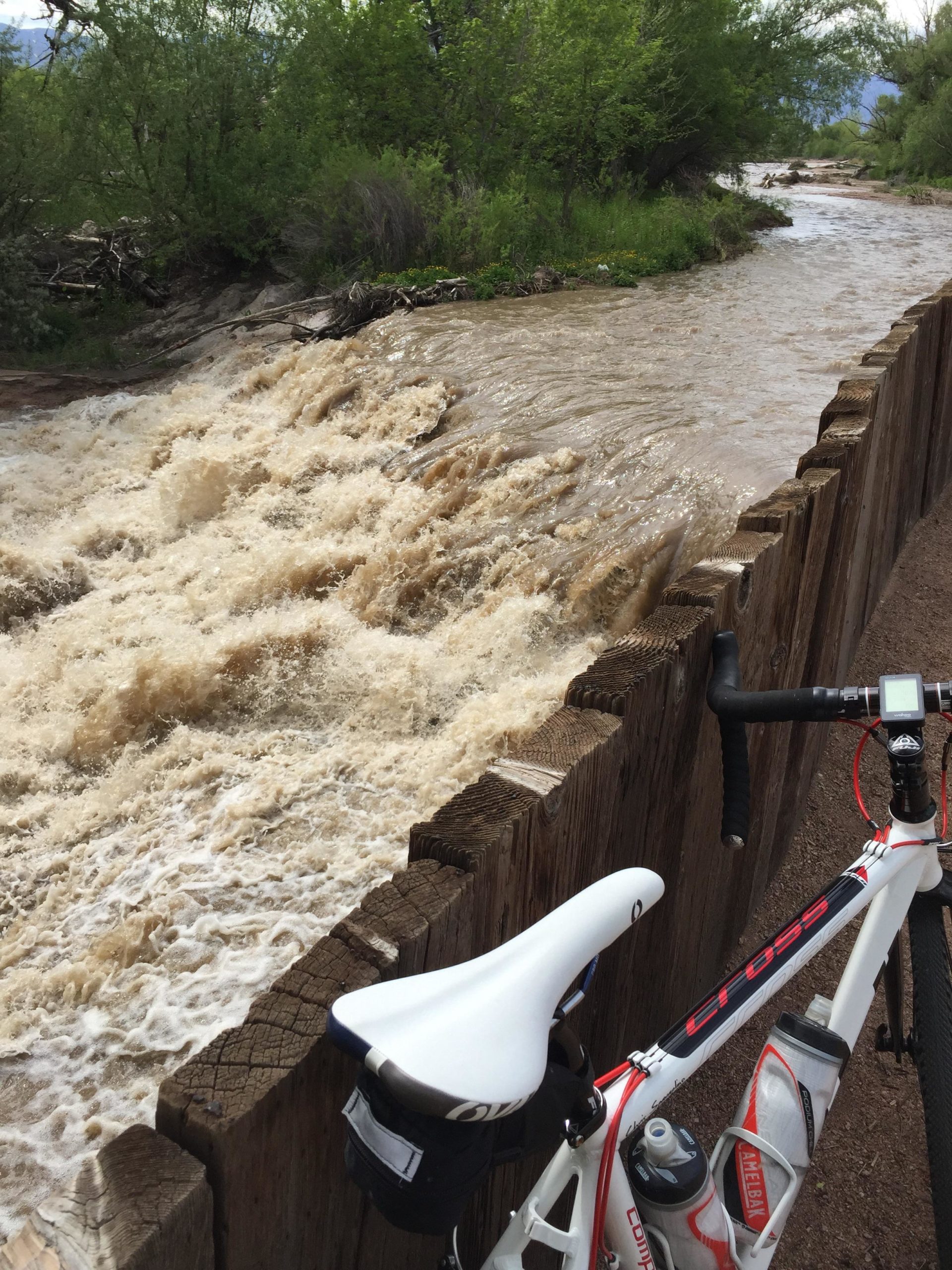 A white and red bicycle rests beside a rapidly flowing river, with brown muddy water and frothy waves. A wooden barrier runs along the edge of the riverbank, surrounded by lush greenery and trees. The scene captures the power of nature and suggests recent heavy rainfall. Santa Fe Trail mountain bike trail.