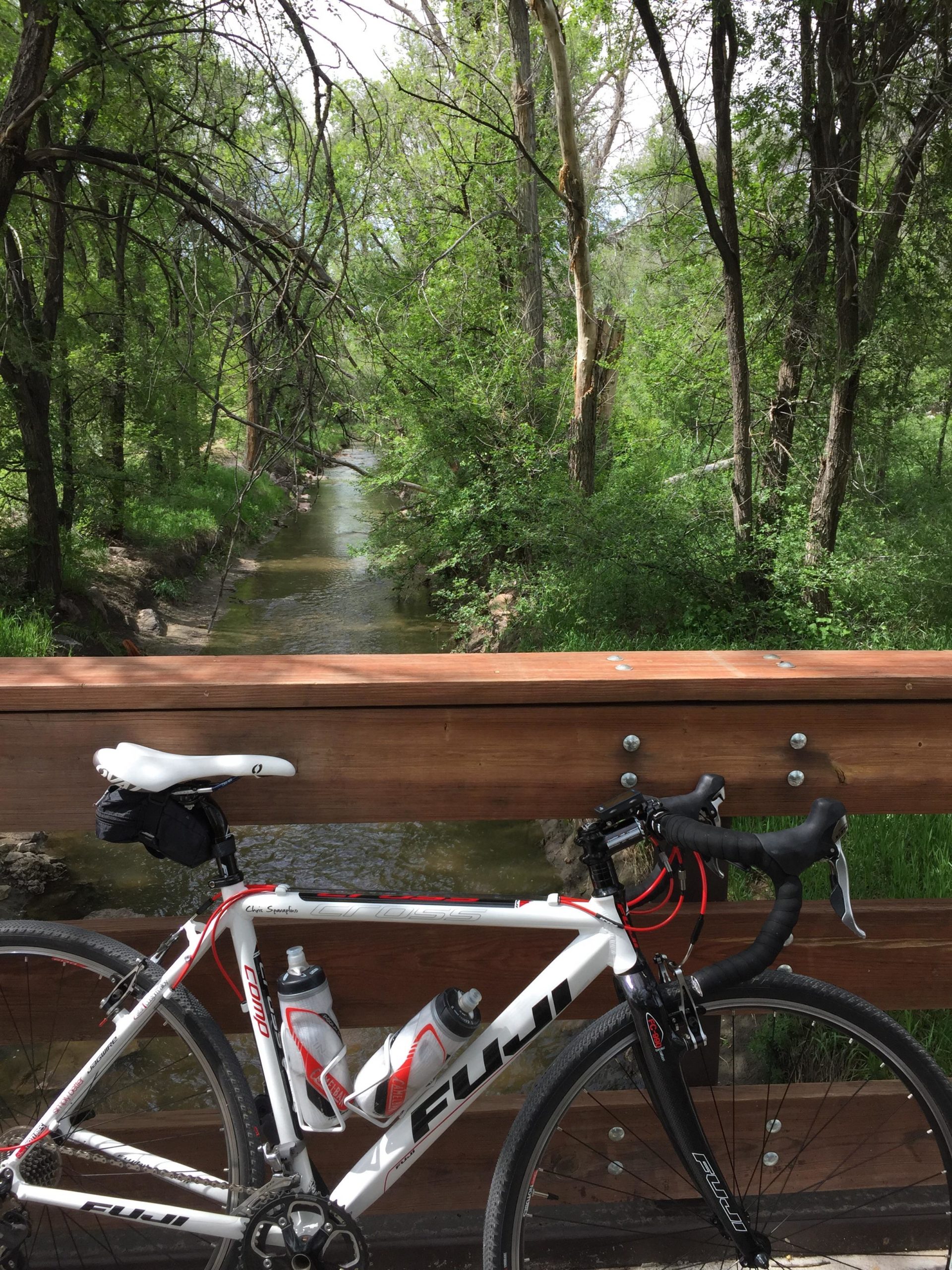 A white Fuji bicycle is parked on a wooden bridge overlooking a serene, green landscape with a small creek flowing below. The scene is surrounded by lush trees and foliage, creating a tranquil natural setting on a sunny day. Santa Fe Trail mountain bike trail.