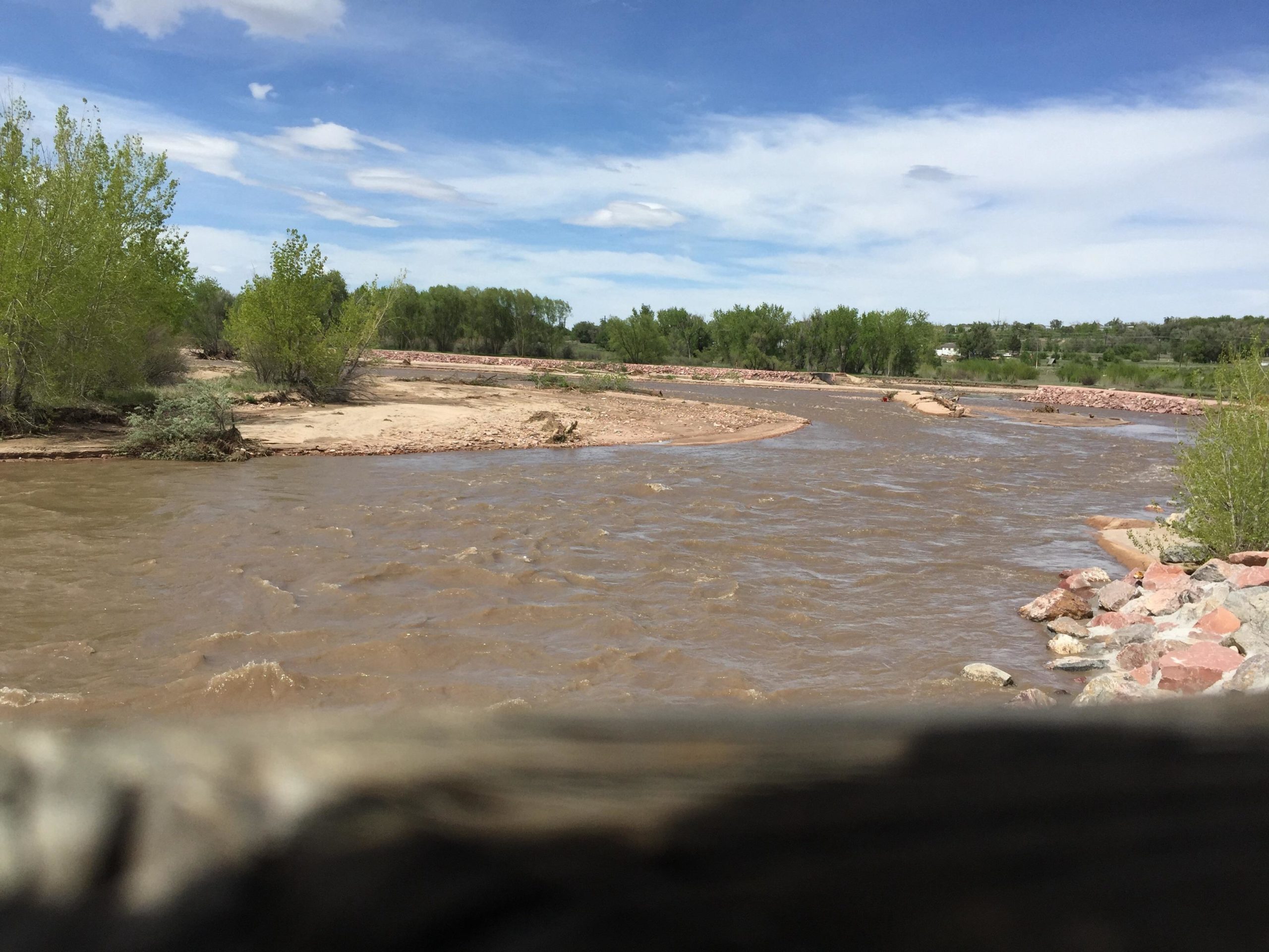 A winding river with muddy water flows through a landscape featuring sandy banks and lush green shrubs. The sky above is partly cloudy, creating a serene outdoor scene. Santa Fe Trail mountain bike trail.