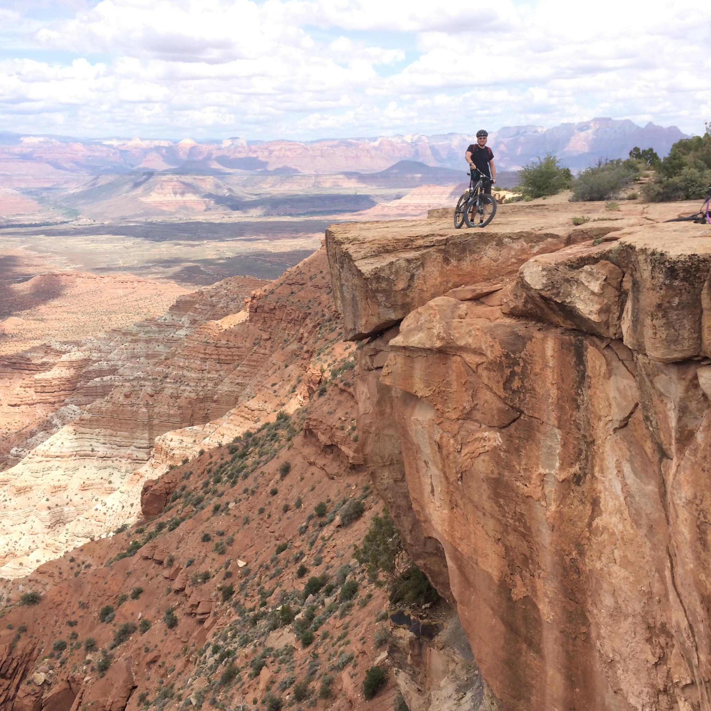 A mountain biker stands near the edge of a rocky cliff overlooking a vast landscape of red rock formations and rolling hills under a cloudy sky. Gooseberry Mesa mountain bike trail.