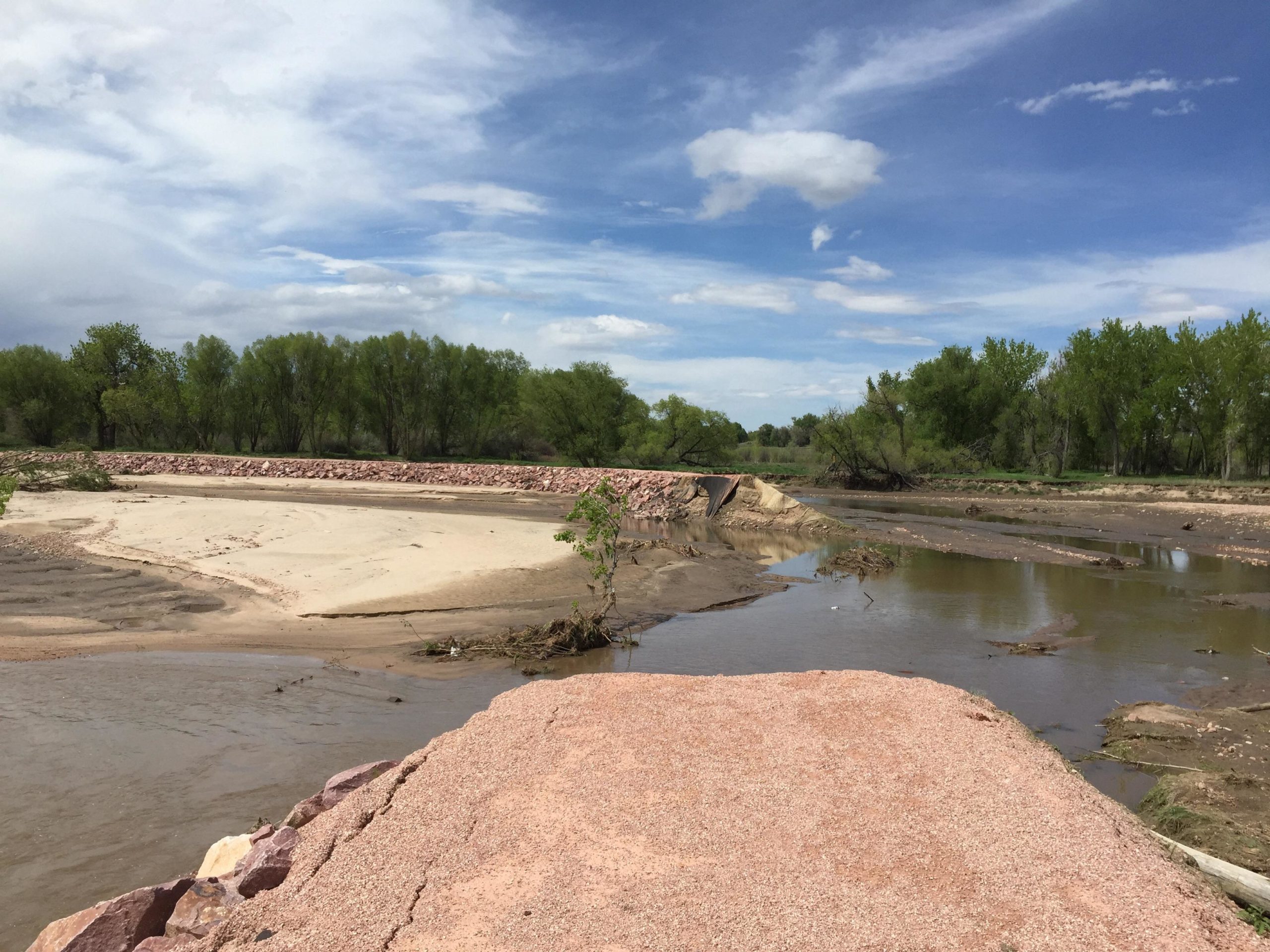 A scenic view of a riverbank with a mixture of sandy and muddy areas, flanked by green trees on the horizon under a partly cloudy blue sky. The river meanders through the landscape, creating a serene natural setting. Santa Fe Trail mountain bike trail.