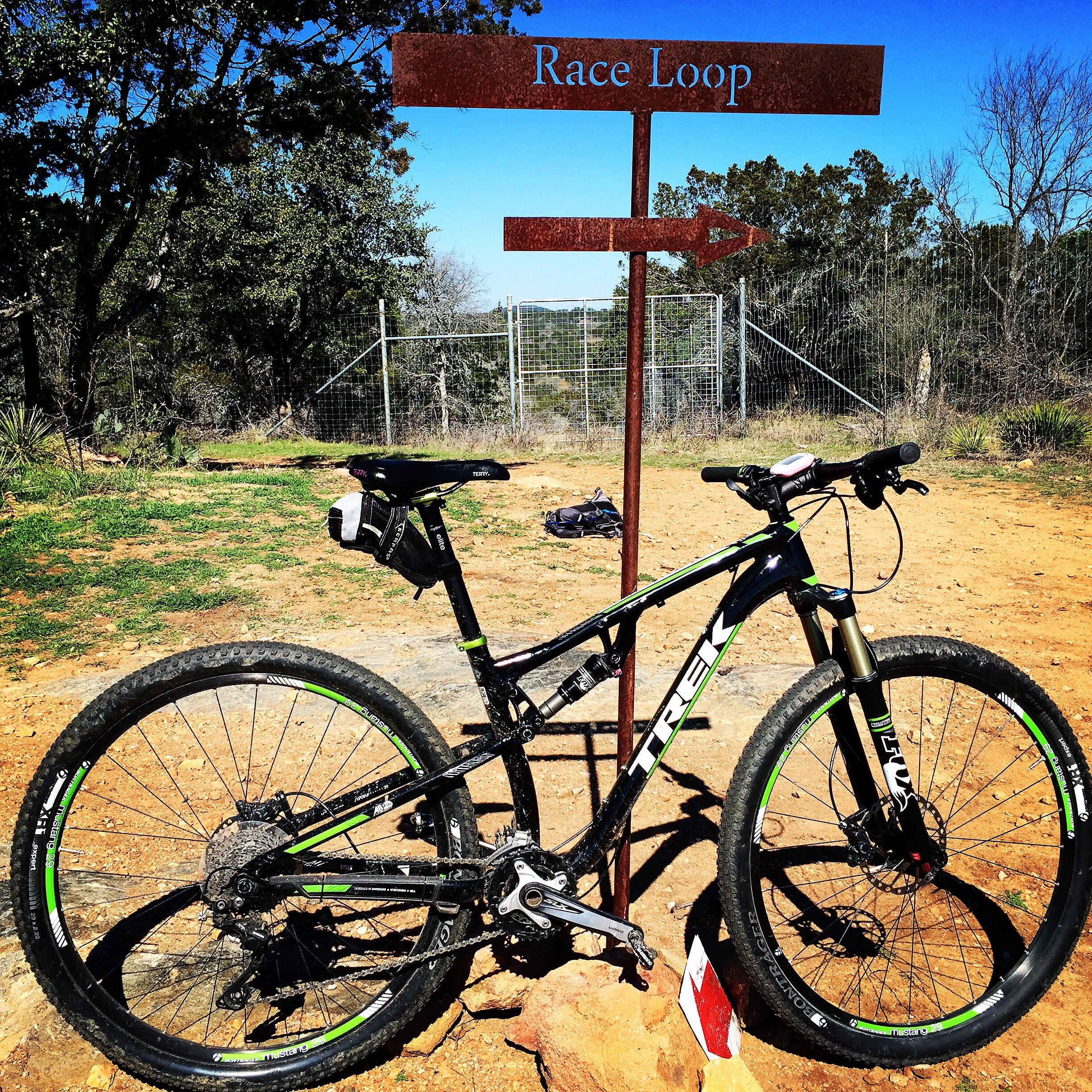 Trek Superfly FS 8: A mountain bike parked beside a signpost labeled "Race Loop," with a clear blue sky and trees in the background. The bike features a black frame with green accents and is positioned on a dirt path surrounded by grassy terrain.