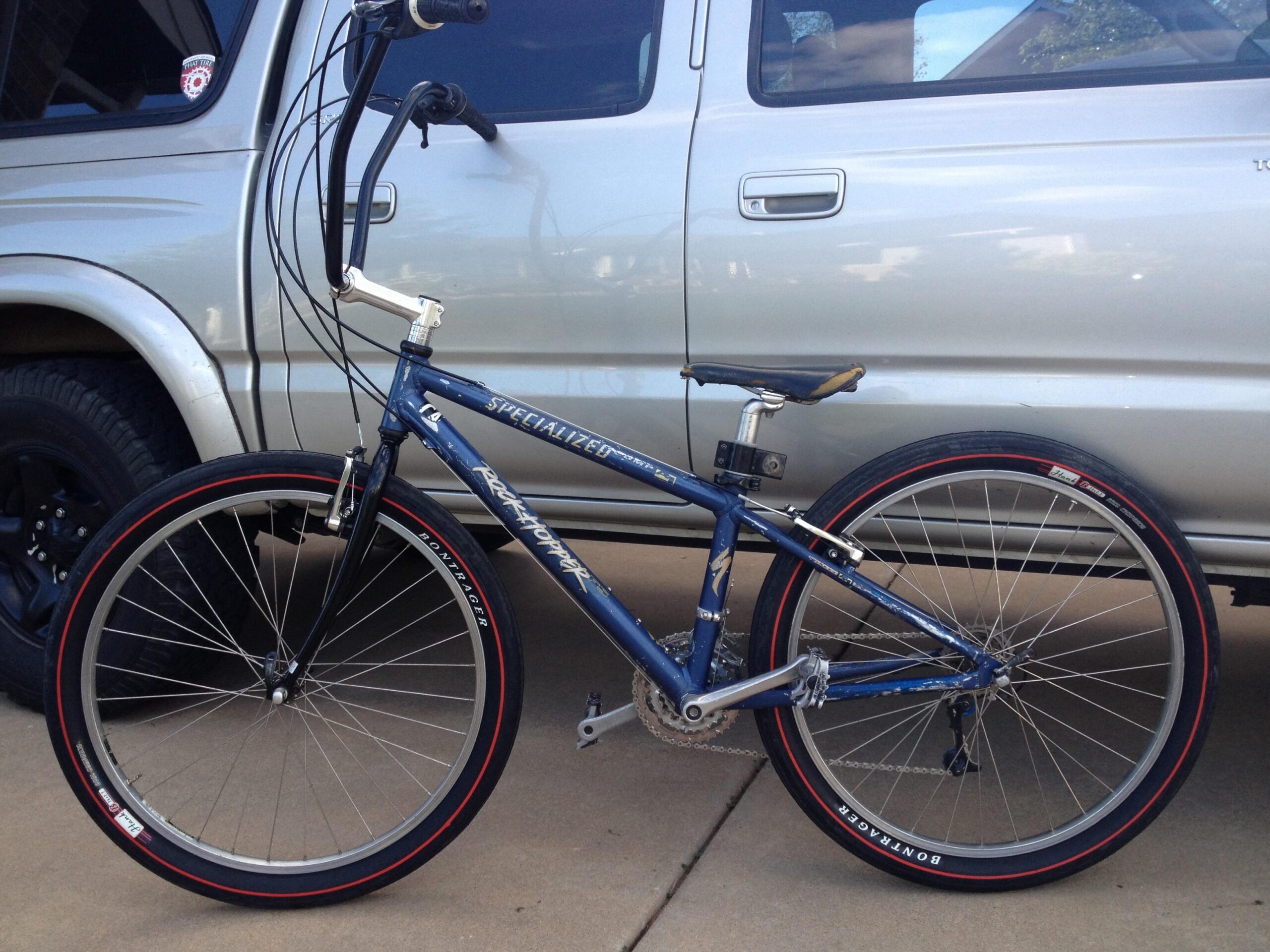 Specialized Rockhopper: A blue Specialized bicycle with distinctive high handlebars and red-trimmed tires parked next to a silver SUV on a concrete surface. The bike features a unique frame design and is positioned prominently in the foreground.