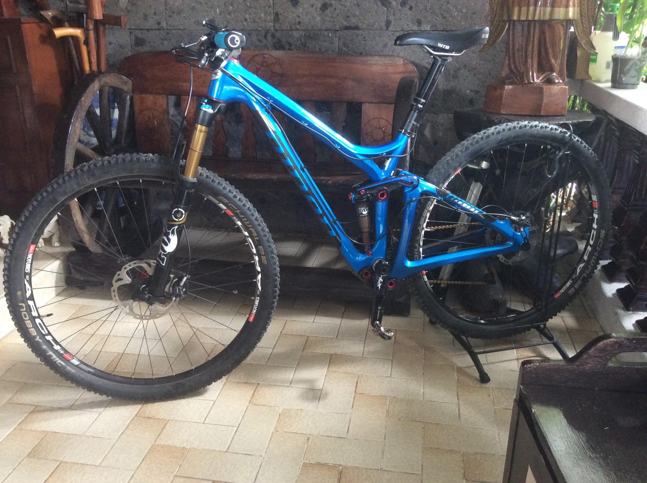 Niner R.I.P. 9 RDO: A blue mountain bike is positioned in a room featuring tiled flooring. The bike has large tires and visible suspension components, with a decorative wooden bench and plant in the background. The environment suggests a cozy indoor setting.