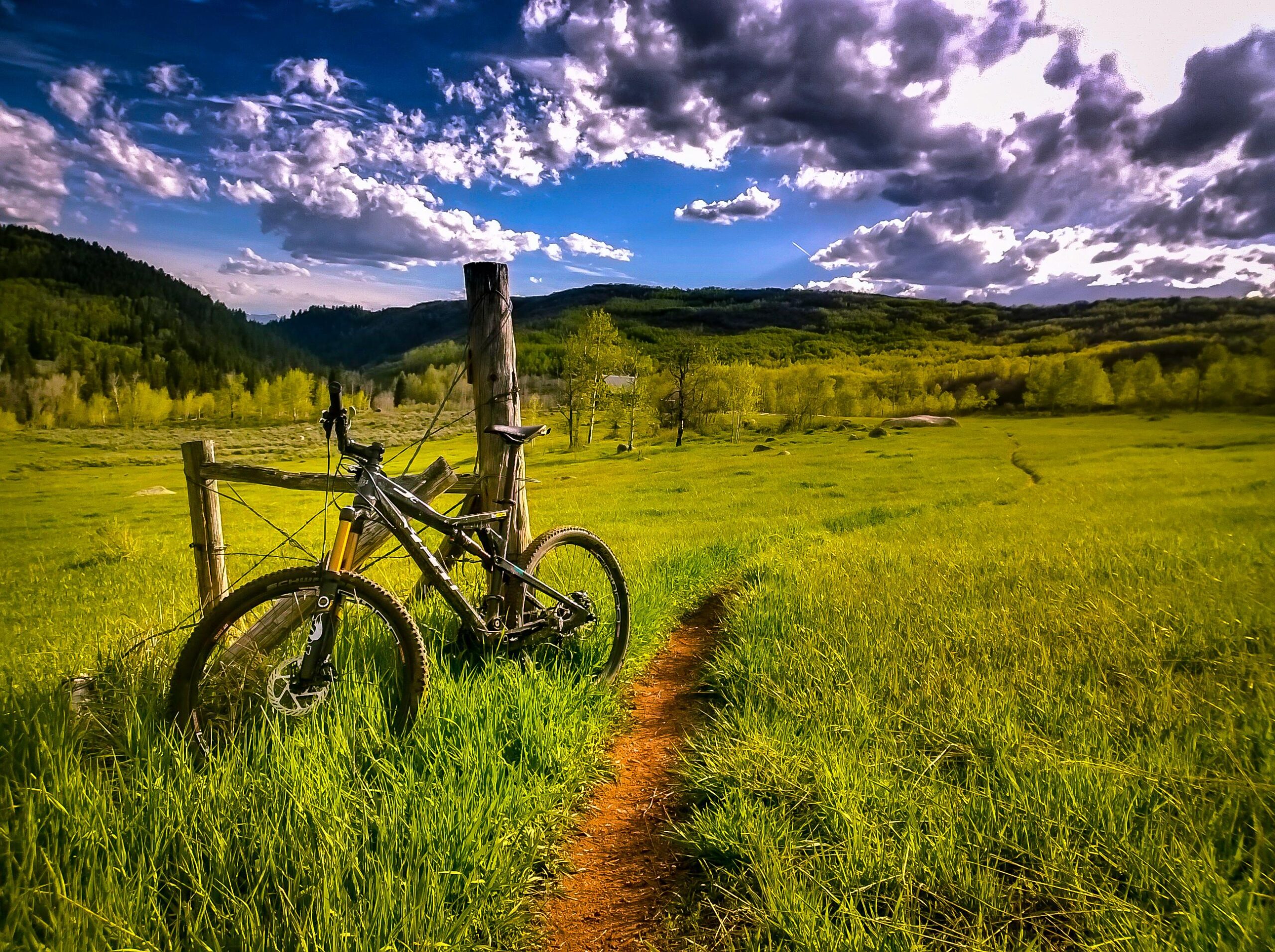 Mountain bike resting against a wooden post on a lush green field, with a winding dirt path leading into the distance. The background features rolling hills and a partly cloudy blue sky. Mad Creek mountain bike trail.