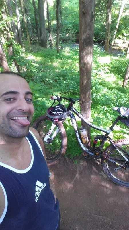 A man sticking out his tongue and smiling in a forested area, with a mountain bike parked next to him. The background features lush green foliage and trees, suggesting an outdoor recreational setting. Six Mile Run mountain bike trail.