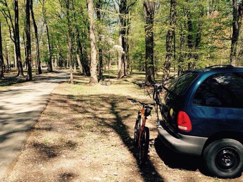 A scenic park area featuring a dirt path winding through trees with vibrant green leaves. An orange bicycle is parked next to a blue minivan, both situated on the grassy edge of the path. In the background, more trees and a hint of another bike can be seen. The image captures a peaceful outdoor atmosphere, ideal for biking or enjoying nature. Freedom Center mountain bike trail.