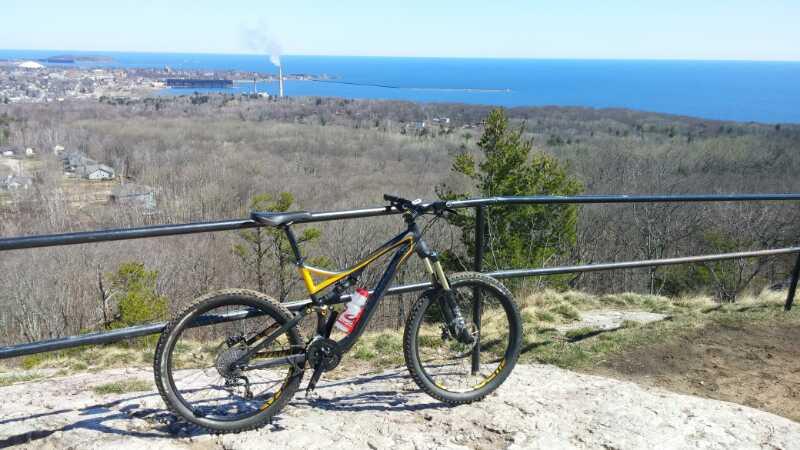 A mountain bike parked on a rocky outcrop overlooking a panoramic view of a lake and distant shore, with trees and a clear blue sky in the background. Noquemanon Trails Network: South Marquette Trails mountain bike trail.