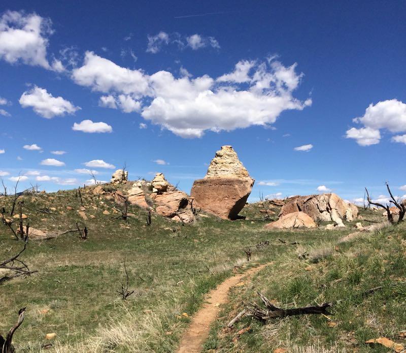 A scenic landscape featuring large rock formations and a clear blue sky with scattered white clouds. In the foreground, a winding dirt path leads through a grassy area interspersed with dry vegetation and fallen branches. The rocky outcrops vary in size and shape, creating a natural and rugged terrain. Oil Well Flats mountain bike trail.