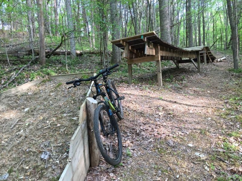 A mountain bike leaning against a wooden ramp in a wooded area, surrounded by trees and fallen leaves. The ramp is designed for bike stunts or tricks, leading into a dirt path. Green River mountain bike trail.