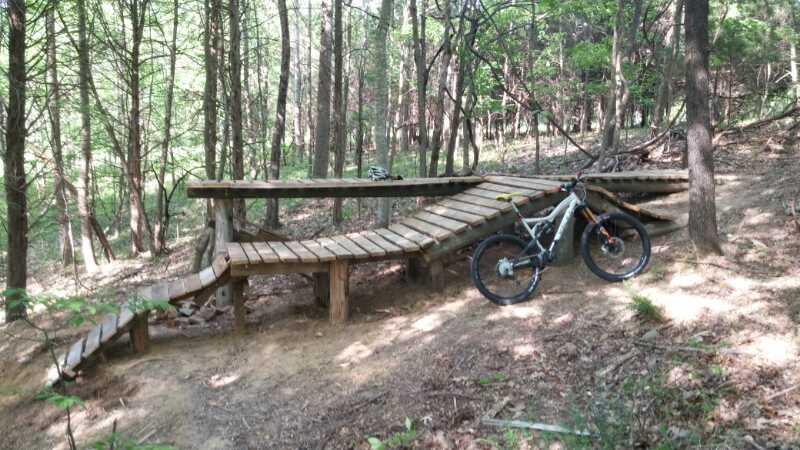 A wooden bike ramp winding through a forested area, designed for mountain biking. The ramp features gradual inclines and declines, with a mountain bike parked beside it on the trail, surrounded by trees and greenery. Green River mountain bike trail.