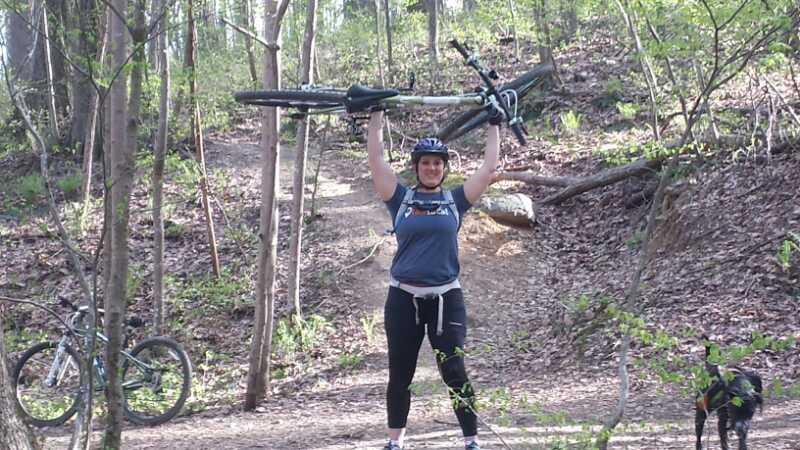 A person wearing a helmet and cycling gear proudly lifts a mountain bike above their head while standing on a dirt trail in a wooded area. A dog is nearby, and another bike is leaning against a tree in the background. Fairland Recreational Park mountain bike trail.