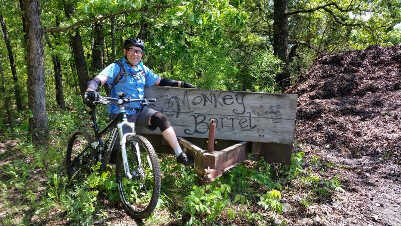 A cyclist in a blue jersey and helmet poses next to a wooden sign that reads "Monkey Barret" while leaning on their mountain bike. The background features lush green trees and a pile of mulch, indicating a natural outdoor setting. Forest City Trail mountain bike trail.
