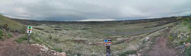 A panoramic view of a grassy landscape with rolling hills under a cloudy sky. In the foreground, there's a dirt path leading to two trail markers: one with a green circle and an arrow pointing left, and another featuring various trail symbols. The scene conveys a sense of outdoor exploration and adventure. Rhyolite Bike Park mountain bike trail.