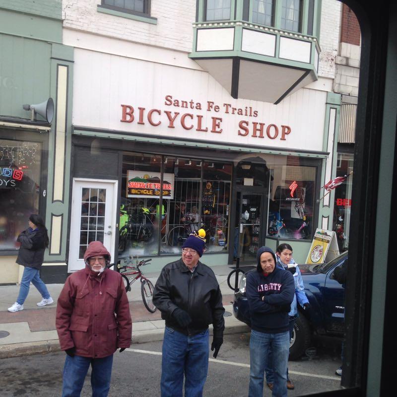 A busy street scene featuring the Santa Fe Trails Bicycle Shop, showcasing its sign prominently. In the foreground, three men are standing on the sidewalk, with a backdrop of a bustling town. One man wears a dark coat and a hat, another has a lighter jacket, and the third stands with crossed arms. Bicycles are visible in the shop window, and passersby are seen in the background. The weather appears cool, suggesting a lively urban atmosphere.