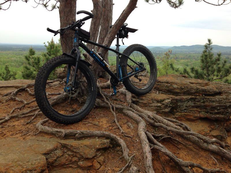 A mountain bike leaning against a tree, positioned on rocky terrain with visible tree roots, overlooking a lush green landscape and distant hills in the background. The scene is set under a cloudy sky. Levis Mounds mountain bike trail.