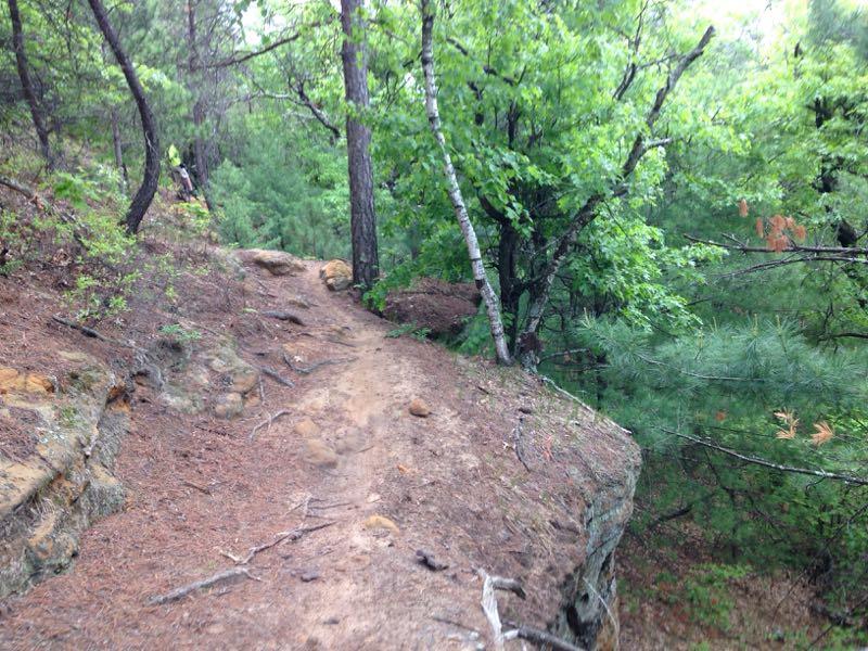 A narrow dirt path winding along a rocky ledge, surrounded by lush green trees and underbrush, indicating a natural hiking trail in a forested area. Levis Mounds mountain bike trail.