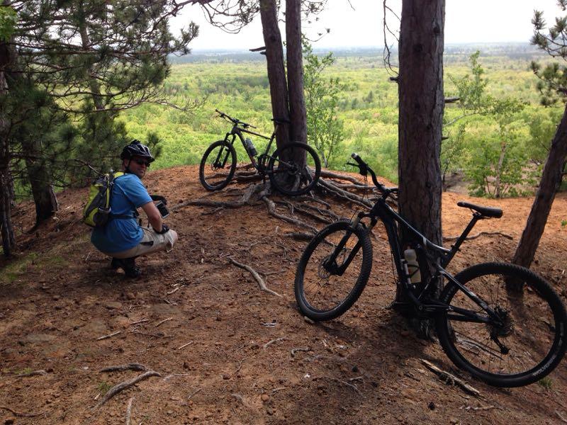 A person in a blue shirt and helmet crouches on a sandy trail surrounded by pine trees, with two mountain bikes resting nearby. The scene overlooks a lush, green landscape in the background under a partly cloudy sky. Levis Mounds mountain bike trail.
