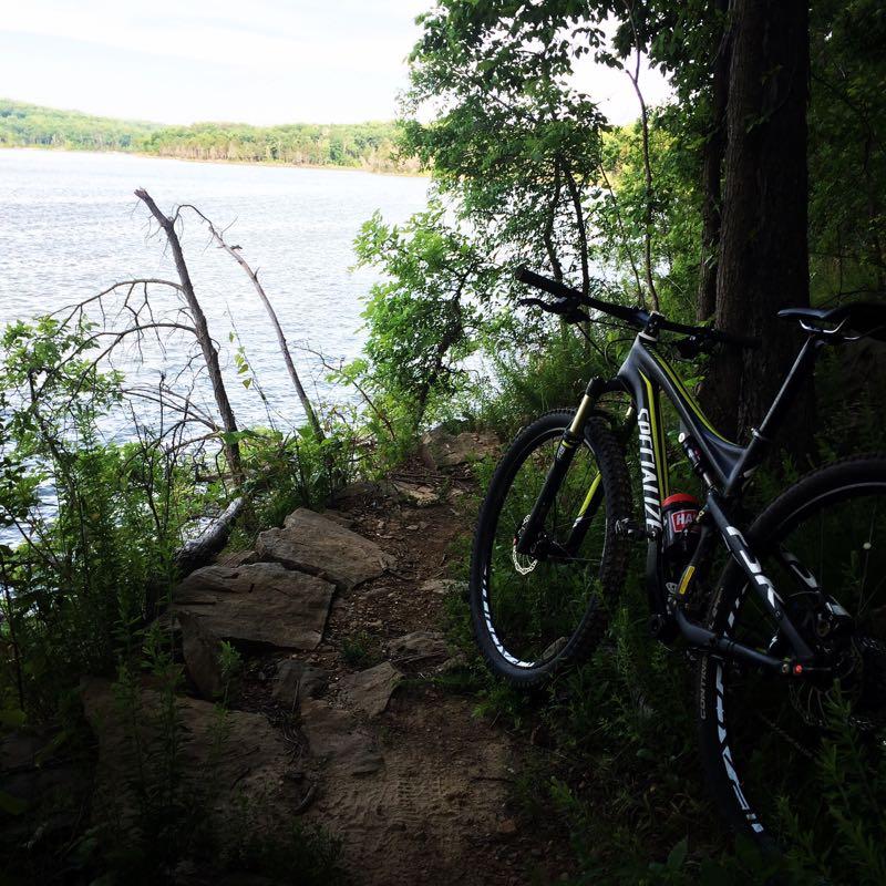 A mountain bike is positioned beside a narrow dirt path leading to a serene lake, surrounded by lush greenery and trees. The water reflects the sky, creating a peaceful outdoor scene perfect for biking enthusiasts. Brier Creek mountain bike trail.