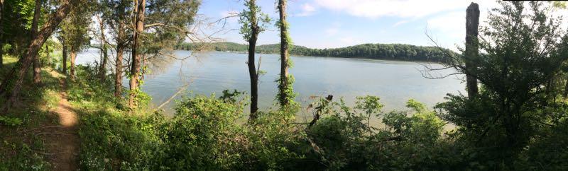 A panoramic view of a serene lake bordered by lush greenery, featuring tall trees and bushes along the shoreline. The calm water reflects the blue sky and distant hills, creating a peaceful natural landscape. Brier Creek mountain bike trail.
