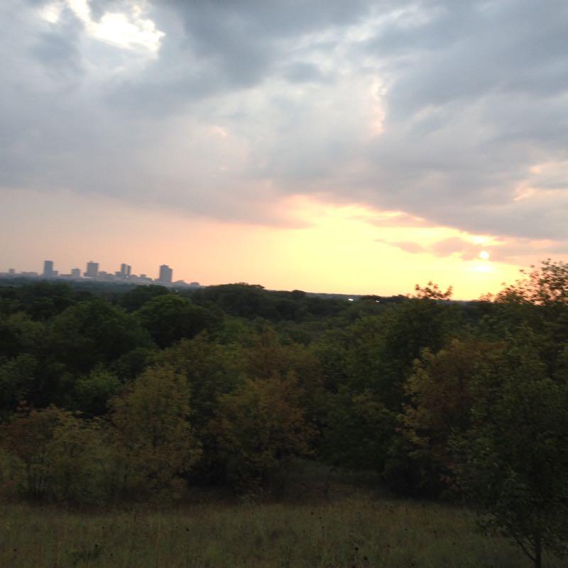 A scenic view of a city skyline silhouetted against a colorful sunset, with lush green trees in the foreground and a cloudy sky creating a dramatic atmosphere. Gateway Park mountain bike trail.