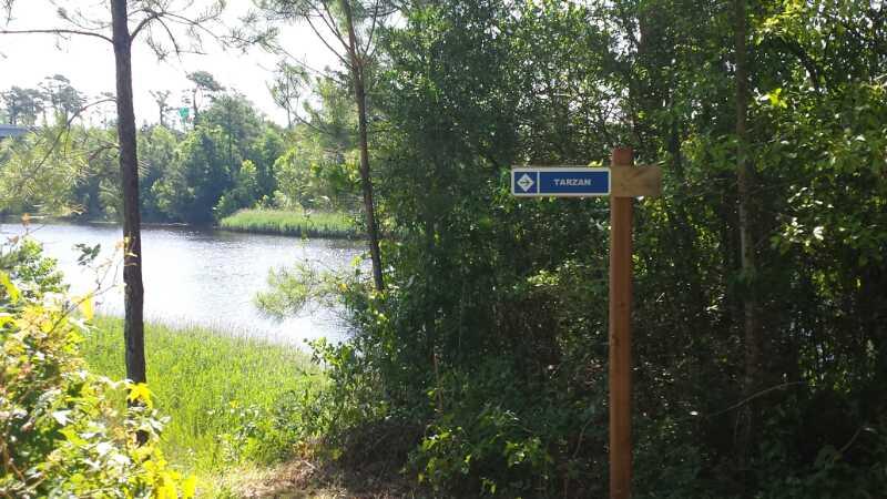 Signpost indicating the "Tarzan" trail, surrounded by lush greenery and near a calm body of water under bright daylight. Horry County Bike Run Park mountain bike trail.