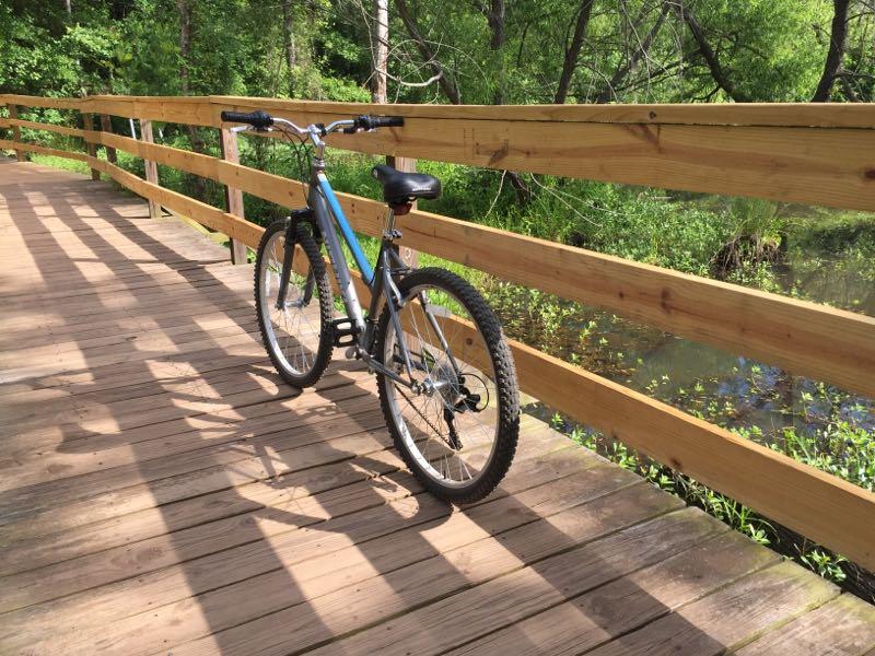 A blue and black mountain bike stands on a wooden bridge, surrounded by lush greenery. The sunlight casts shadows on the deck, and a small body of water is visible nearby. Lake Conestee Nature Park mountain bike trail.
