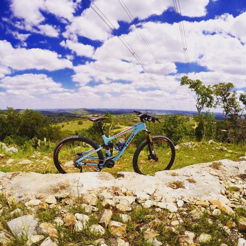 A blue mountain bike rests on a rocky outcrop, with a scenic landscape of rolling green hills and scattered trees in the background. The sky is bright with fluffy white clouds and power lines stretching across the horizon. Flat Rock Ranch mountain bike trail.