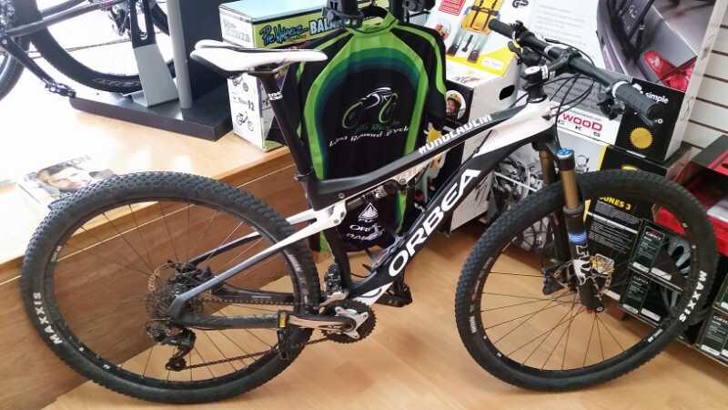 A black and white Orbea mountain bike displayed in a bike shop, featuring knobby tires and a sturdy frame. In the background, a green cycling jersey hangs on a rack, and various cycling accessories and equipment are visible on shelves.