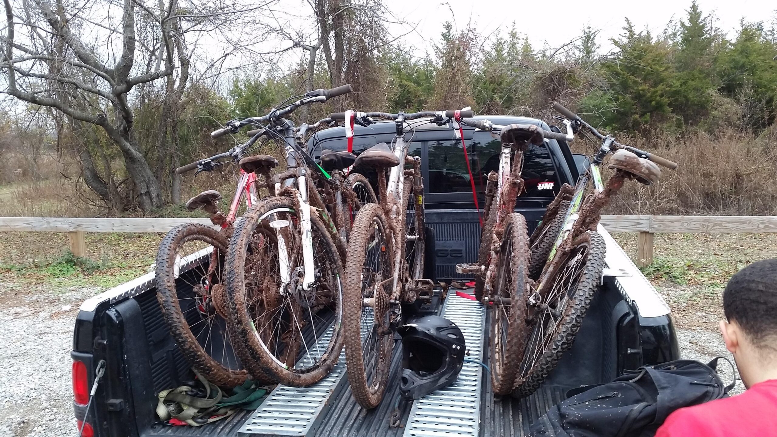A pickup truck bed filled with several muddy mountain bikes, resting against a backdrop of trees. A black bike helmet is placed on the truck bed, and a backpack is partially visible on the right side. The scene captures a post-ride moment, showcasing the equipment used for outdoor biking activities. Six Mile Run mountain bike trail.
