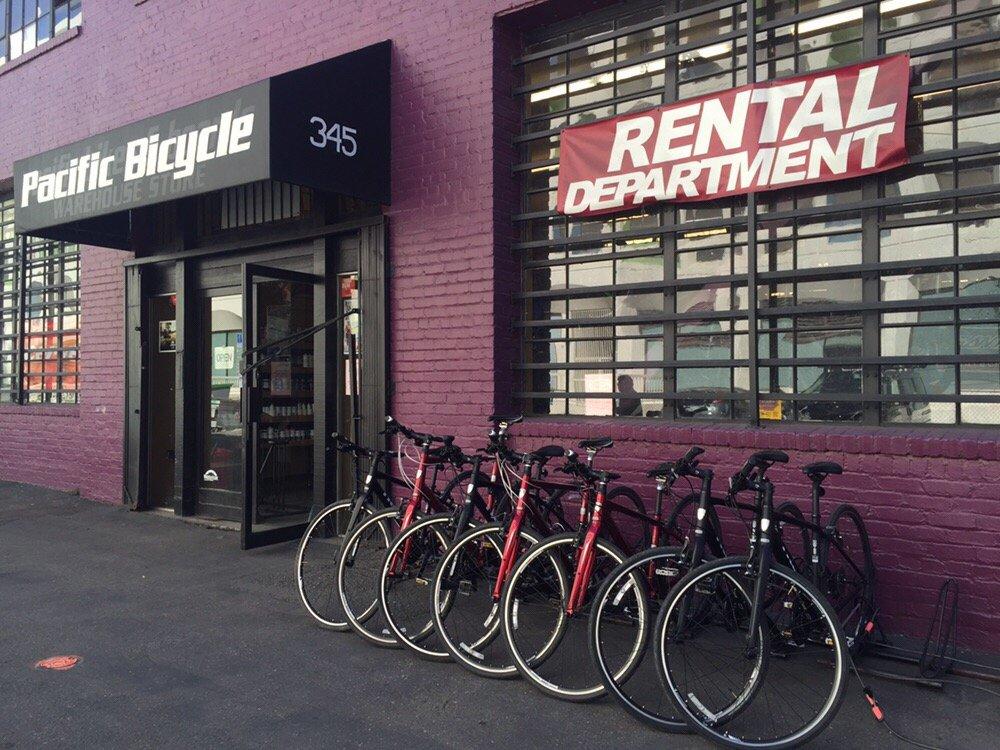 Exterior view of a bicycle rental shop named "Pacific Bicycle," featuring a prominent sign above the entrance. A banner that reads "RENTAL DEPARTMENT" hangs in the window. Several bicycles are neatly parked in front of the store, showcasing different styles and colors against a purple brick wall.
