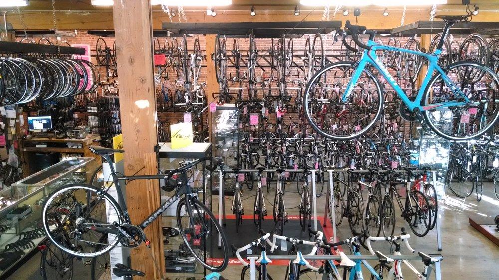 A bicycle shop interior showcasing a variety of bicycles hanging on walls and displayed on racks. Several black road bikes are prominently featured in the foreground, with a bright blue bike suspended above them. The store