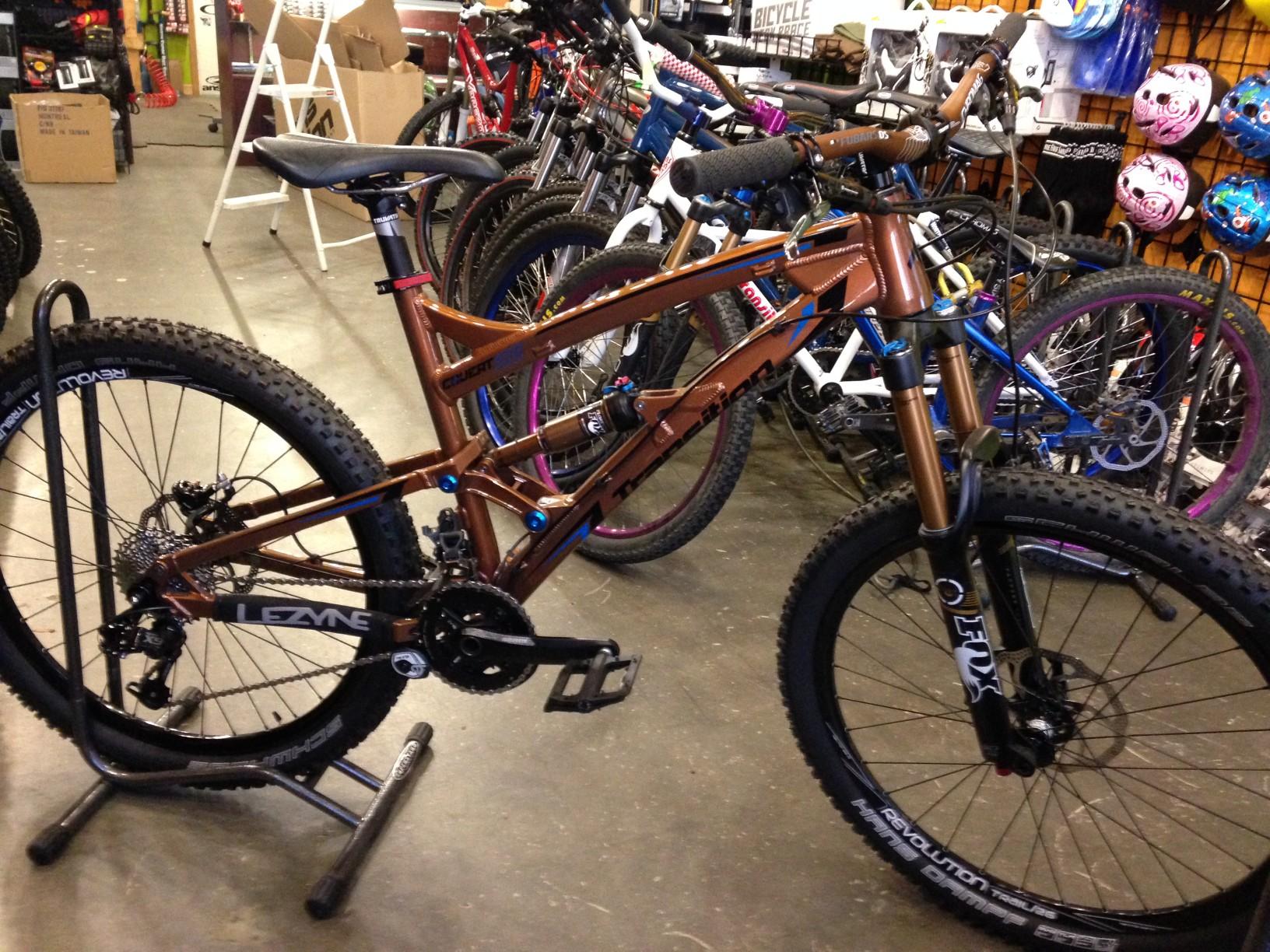 Transition Covert: A mountain bike with a matte brown frame, featuring front suspension and wide, knobby tires. The bike is displayed on a stand in a bike shop, with various other bikes and helmets visible in the background.