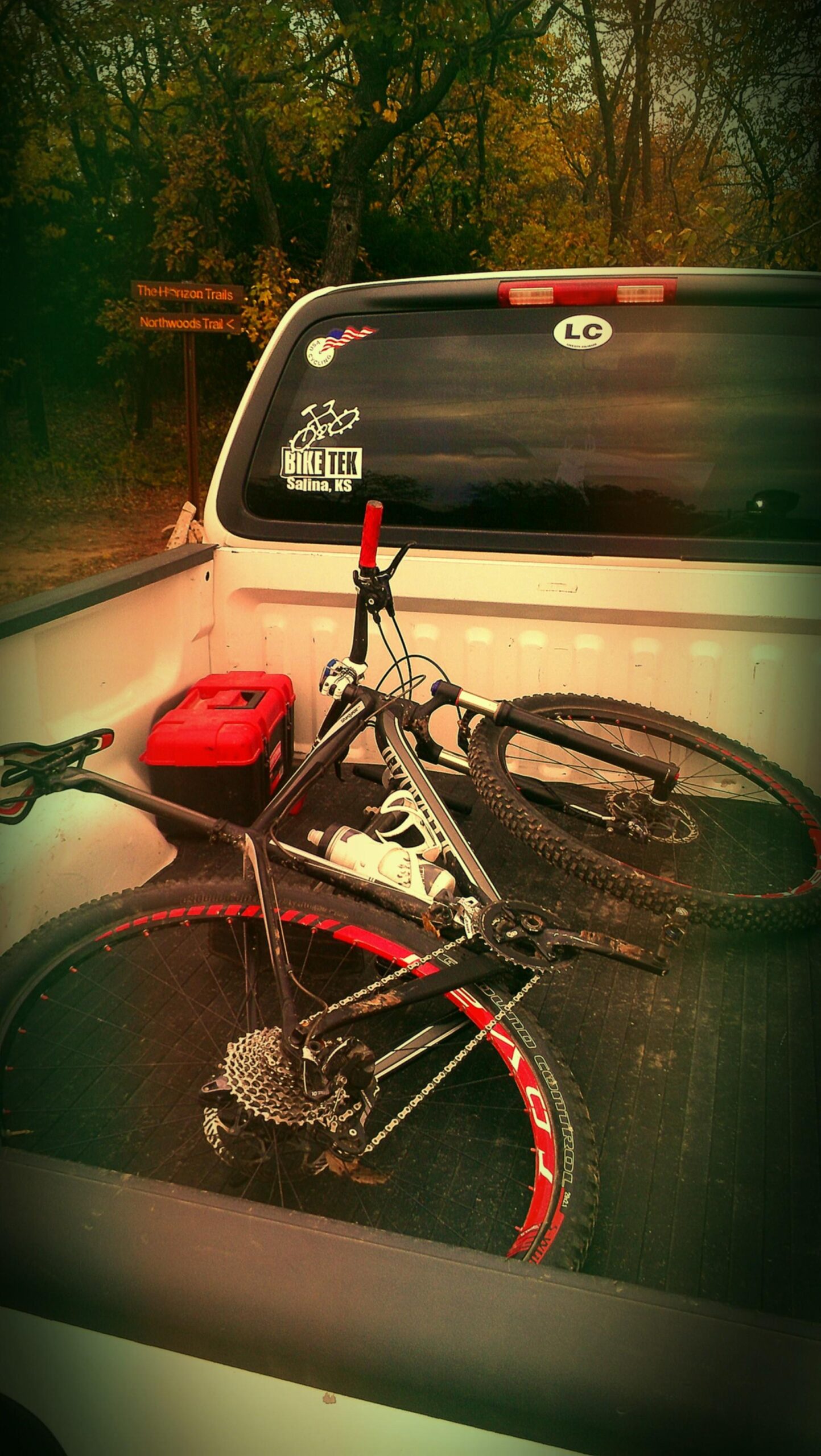 A close-up view of a pickup truck bed containing two mountain bikes with red accents, a water bottle, and a red toolbox. In the background, a wooden signpost indicates trail names. The setting is surrounded by trees with autumn foliage, giving the scene a rustic outdoor feel. Camp Horizon mountain bike trail.