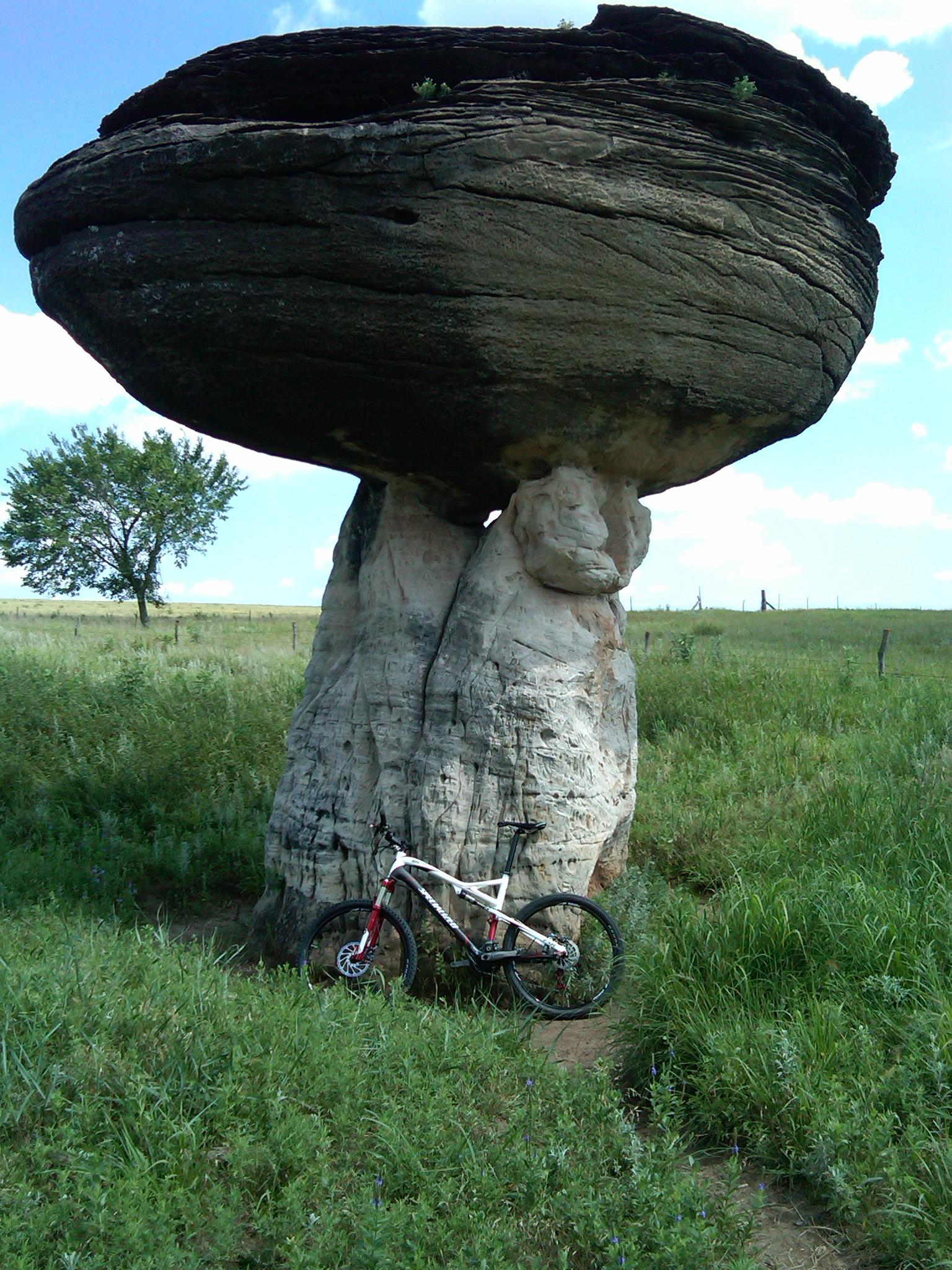 A large, flat-topped rock formation resembling a mushroom stands in a grassy field, with a mountain bike leaning against its base. In the background, a single tree and a blue sky with scattered clouds are visible. Kanopolis Lake mountain bike trail.