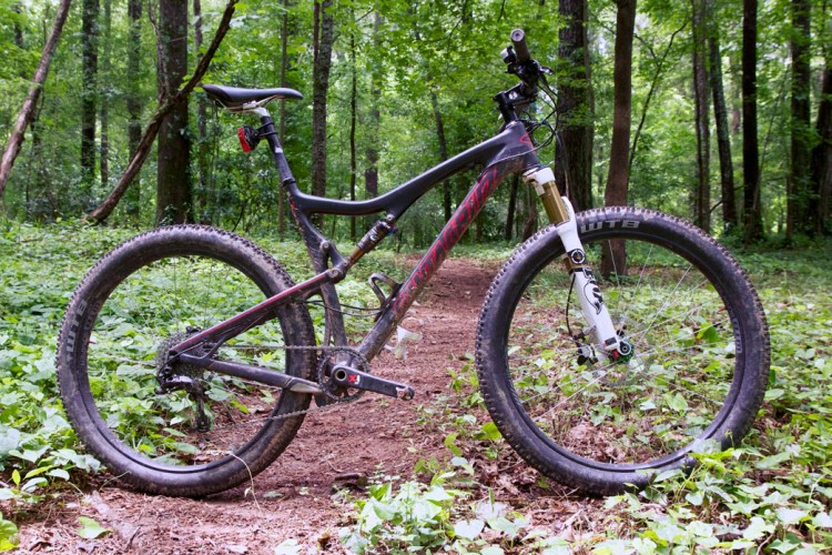 A mountain bike parked on a trail surrounded by lush green foliage in a wooded area. The bike features large tires and a sturdy frame, suggesting it is designed for off-road riding.