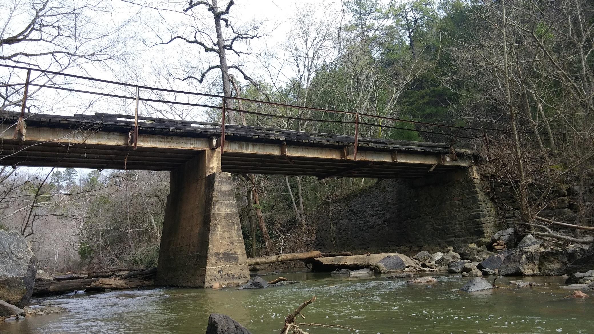 Old bridge spanning a calm river, supported by stone and concrete pillars, with trees and rocky banks lining the waterway. The bridge features a weathered wooden deck and rusted railing, set against a backdrop of bare trees and lush greenery in the surrounding landscape. Chewacla State Park mountain bike trail.